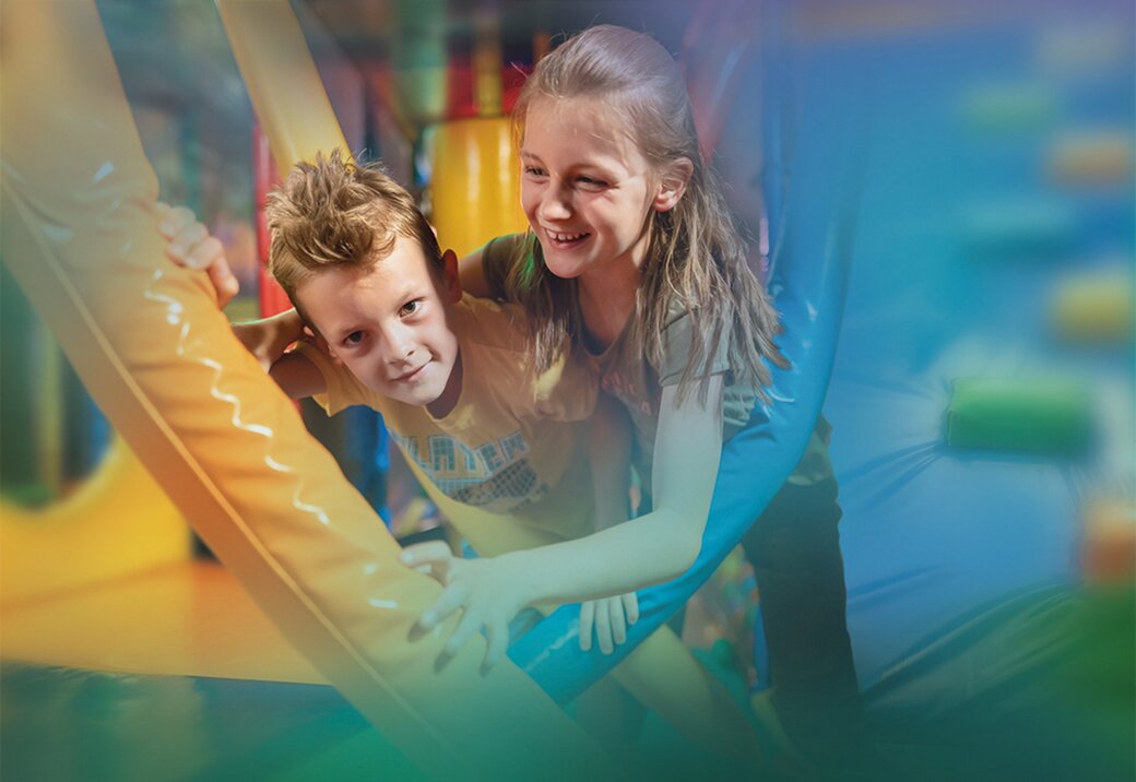 Zwei Kinder spielen und lachen auf einer blauen Rutsche in einem bunten Indoorspielplatz.