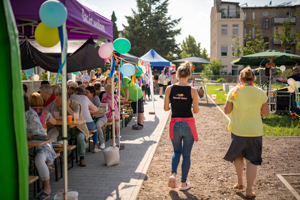 Bunt geschmücktes Straßenfest: Viele Menschen sitzen unter Pavillons an langen Tischen, während zwei Helferinnen über den Weg Richtung Bühne gehen.