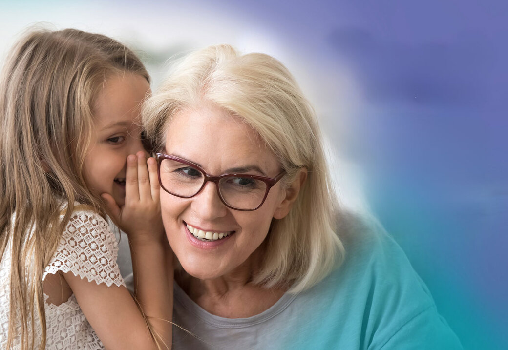 Ein Mädchen flüstert einer Frau mit Brille und blonden Haaren etwas in ihr Ohr. Beide lächeln dabei.