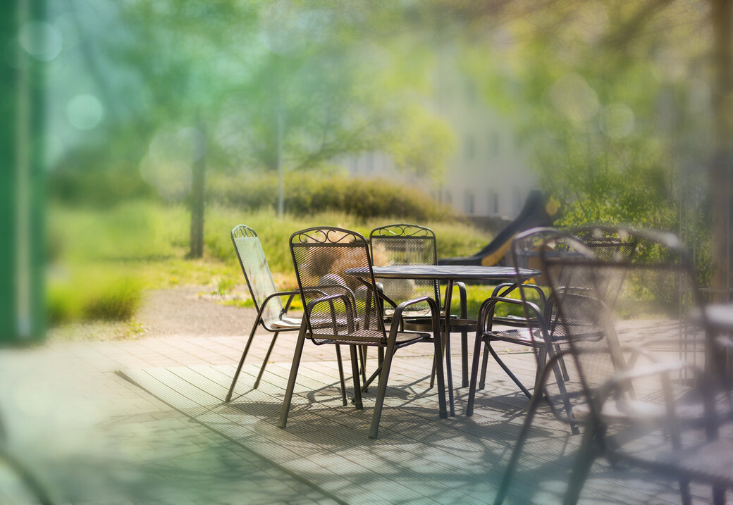 Sonnige Terrasse mit Metalltisch und Stühlen im Grünen, weich unscharfer Hintergrund mit Bäumen und Wohnhaus