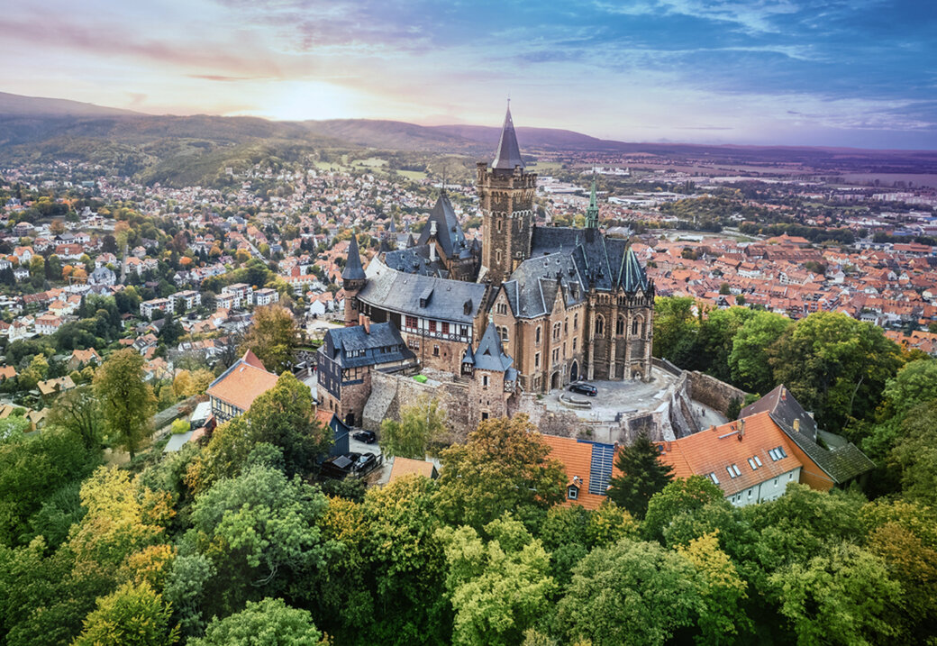 Luftaufnahme eines Schlosses auf einem bewaldeten Hügel, dahinter eine weitläufige Stadt und sanfte Hügellandschaft im Abendlicht.